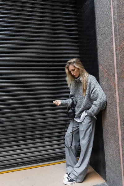 Comprimento total da mulher loira em roupas de inverno em pé com a mão no bolso e segurando bolsa ao ar livre — Fotografia de Stock