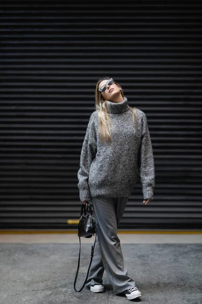 Comprimento total de mulher loira em camisola de inverno e óculos de sol andando com bolsa na rua urbana — Fotografia de Stock