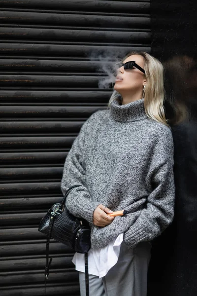 Mulher loira em camisola de inverno e óculos de sol fumando enquanto está de pé com bolsa na rua urbana — Fotografia de Stock