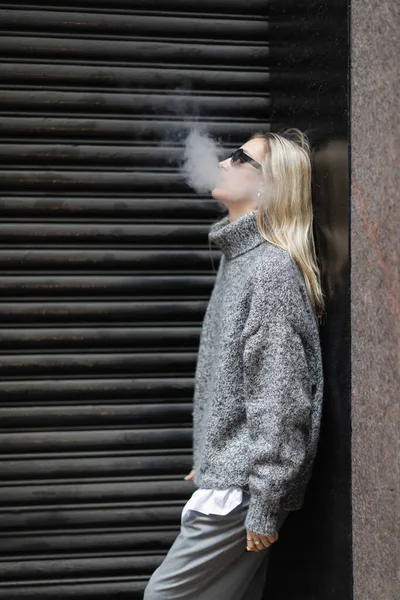 Vista lateral de la mujer joven en suéter de invierno y gafas de sol fumar cigarrillo electrónico mientras está de pie en la calle urbana - foto de stock