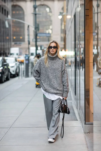 Comprimento total de mulher loira em camisola de inverno e óculos de sol andando com bolsa na rua urbana em Nova York — Fotografia de Stock