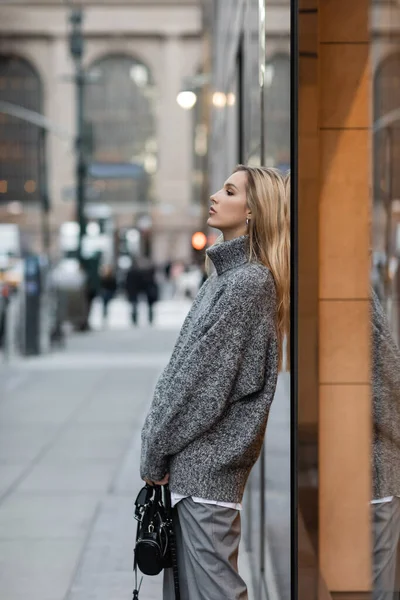 Vista lateral da mulher loira em camisola de inverno em pé com bolsa na rua urbana em Nova York — Fotografia de Stock