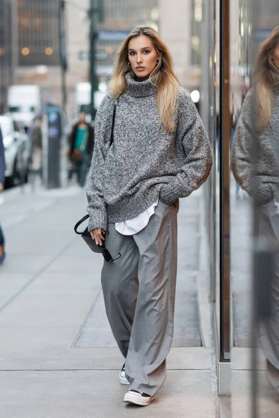 Comprimento total de mulher loira em camisola de inverno segurando óculos de sol e bolsa na rua urbana em Nova York — Fotografia de Stock