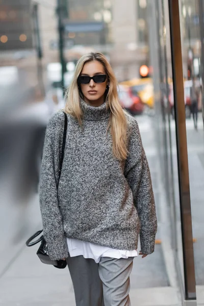Mulher loira em camisola de inverno e óculos de sol andando com bolsa na rua urbana na cidade de Nova York — Fotografia de Stock
