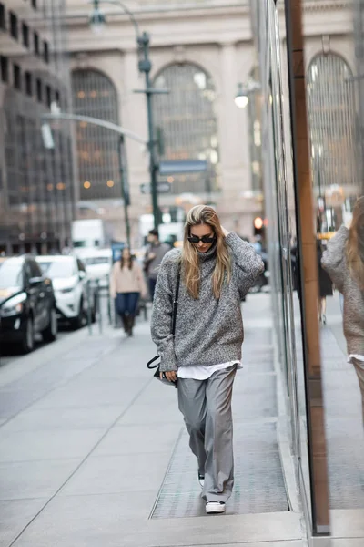 Comprimento total da mulher em camisola de inverno e óculos de sol andando com bolsa na rua urbana em Nova York — Fotografia de Stock