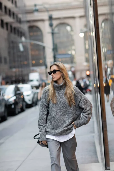 Bonita mulher em camisola de inverno e óculos de sol de pé com a mão no quadril na rua urbana em Nova York — Fotografia de Stock