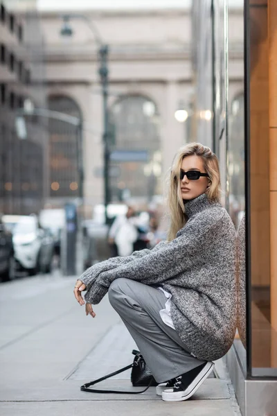 Молода жінка в зимовому светрі й сонцезахисні окуляри сидять біля сумочки на міській вулиці в місті Нью - Йорку. — стокове фото