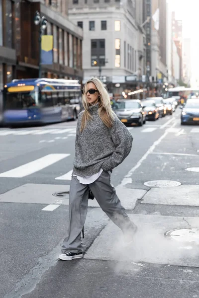 Full length of blonde woman in stylish winter sweater and sunglasses walking on urban street in New York — Stock Photo
