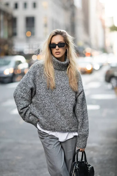 Блондинка в трикотажном свитере и солнцезащитных очках стоит с модной сумочкой на размытой улице Нью-Йорка — стоковое фото
