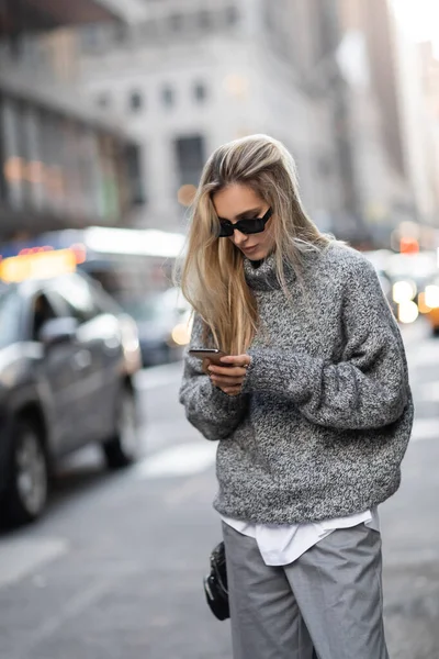 Mulher concentrada em camisola de inverno e óculos de sol usando smartphone na rua urbana na cidade de Nova York — Fotografia de Stock