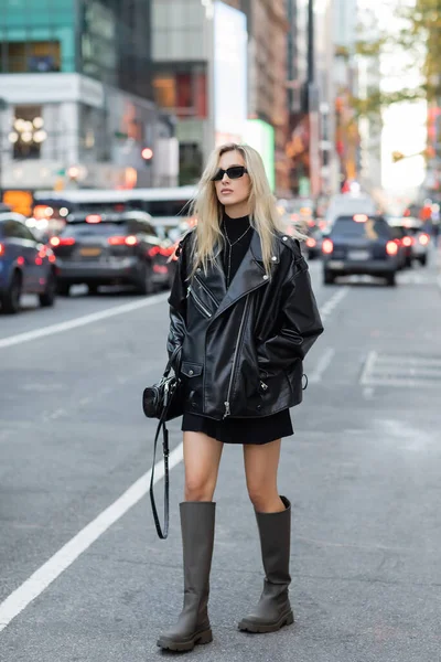Comprimento total de mulher jovem elegante em jaqueta de couro e óculos de sol andando com as mãos em bolsos na rua de Nova York — Fotografia de Stock