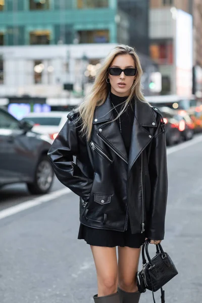 Молода жінка в шкіряній куртці і сонцезахисних окулярах стоїть з рукою в кишені на вулиці Нью-Йорку — стокове фото