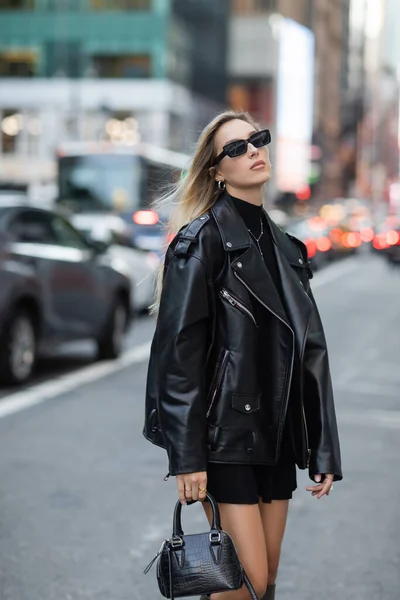 Стильная женщина в кожаной куртке и черном платье стоя с модной сумочкой на улице Нью-Йорка — стоковое фото