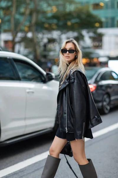 Stylish woman in leather jacket and black dress walking near blurred cars on street of New York — Stock Photo