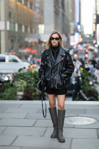 Piena lunghezza di donna elegante in giacca di pelle e vestito nero in piedi con le mani in tasca a New York — Foto stock