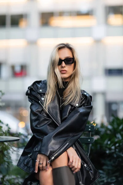 Young blonde woman in black leather jacket and sunglasses sitting in outdoor cafe — Stock Photo