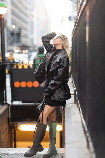 Full length of stylish woman in leather jacket and sunglasses adjusting hair while standing near subway entrance in New York — Stock Photo