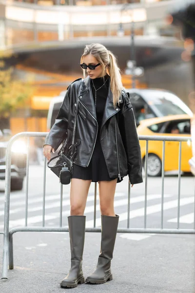 Велика довжина стильної жінки в чорній шкіряній куртці і сукні стоять біля металевого бар'єру в Нью-Йорку — стокове фото