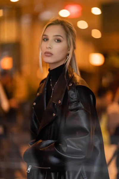 Stylish woman in black leather jacket looking at camera through glass window in New York — Stock Photo