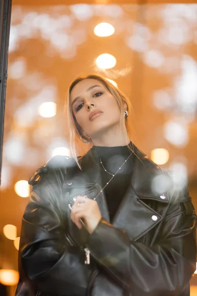 Low angle view of stylish woman in black leather jacket looking at camera through glass window in New York — Stock Photo