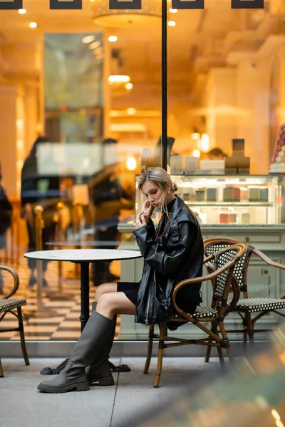 Full length of blonde woman in black leather jacket sitting near round bistro table in outdoor cafe in New York — Stock Photo