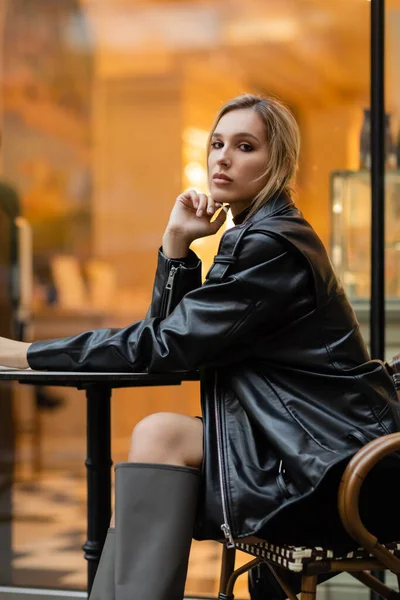Comprimento total da jovem mulher em jaqueta de couro preto sentado perto da mesa redonda bistro no café ao ar livre em Nova York — Fotografia de Stock