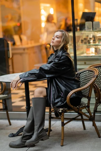 Comprimento total de mulher elegante em jaqueta de couro preto e botas sentadas perto da mesa de bistro no café ao ar livre em Nova York — Fotografia de Stock