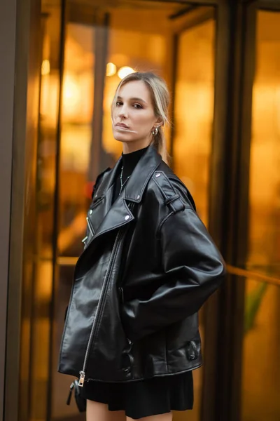 Retrato de mulher jovem e loira em jaqueta de couro preto olhando para a câmera enquanto de pé com a mão no bolso fora — Fotografia de Stock