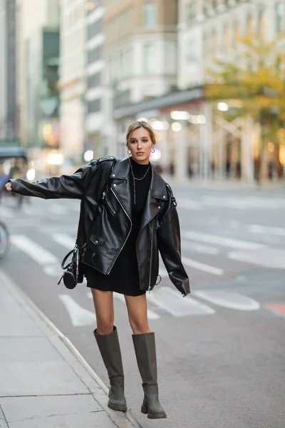 Comprimento total da jovem mulher em jaqueta de couro preto e vestido de pé com a mão estendida na rua em Nova York — Fotografia de Stock