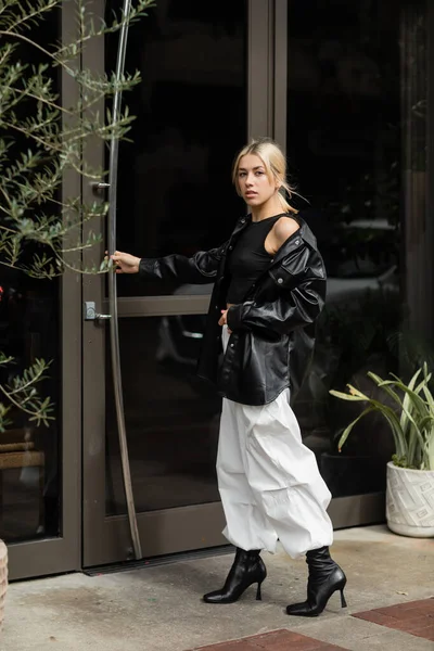 Pleine longueur de femme élégante en veste chemise en cuir et pantalon cargo ouvrant la porte d'entrée sur la rue à Miami — Photo de stock