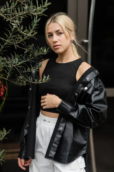 Blonde woman in leather shirt jacket and black tank top standing near green plant — Stock Photo