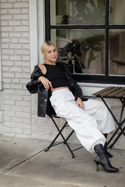 Full length of blonde woman in leather shirt jacket and cargo pants sitting in outdoor cafe on street in Miami — Stock Photo