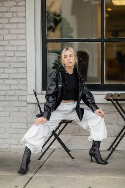 Piena lunghezza di bella donna in giacca camicia di pelle e pantaloni cargo seduto in caffè all'aperto in strada a Miami — Foto stock