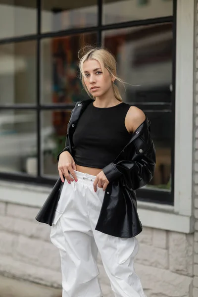 Bonita mujer en chaqueta de camisa de cuero negro y pantalones de carga de pie cerca del edificio en la calle en Miami - foto de stock