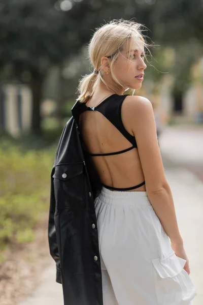 Donna bionda in pantaloni cargo bianchi in piedi con giacca di pelle nera in strada a Miami — Foto stock