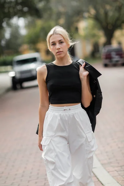 Mujer joven en pantalones de carga blancos y camiseta sin mangas caminando con chaqueta de camisa negra en la calle en Miami - foto de stock