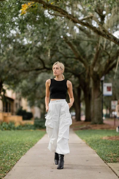Piena lunghezza della donna bionda in pantaloni cargo bianchi e canotta nera che cammina su strada verde a Miami — Foto stock