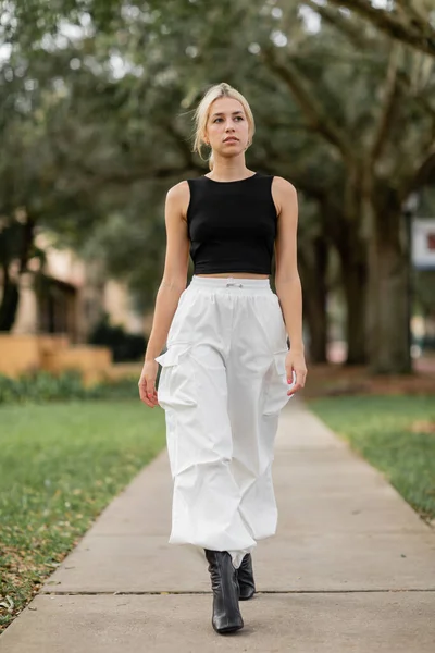 Full length of young woman in white cargo pants and black tank top walking on green street in Miami — Stock Photo