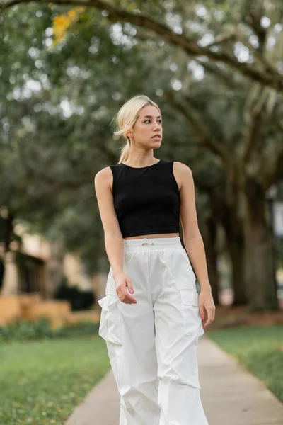 Mujer rubia joven en pantalones de carga blancos y camiseta negra caminando por la calle verde en Miami - foto de stock