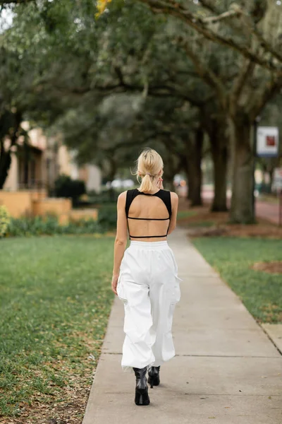 Vista trasera de mujer joven en pantalones de carga blancos y camiseta negra caminando por la calle verde en Miami - foto de stock