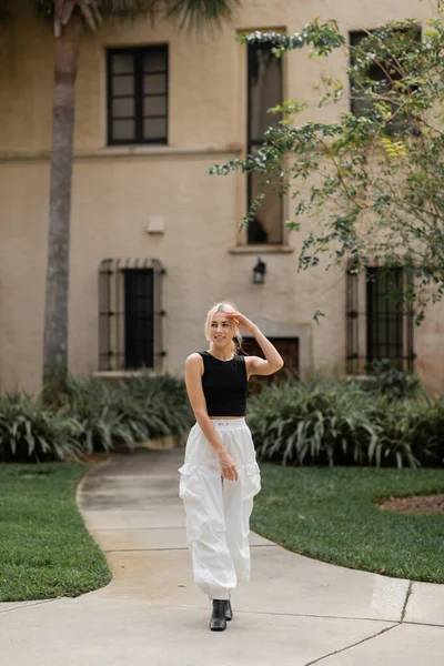 Повна довжина приємної молодої жінки в білих вантажних штанах і модному цистерні, що стоїть біля будинку в Маямі. — Stock Photo