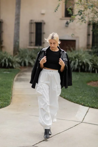 Longitud completa de mujer joven en pantalones de carga blancos y chaqueta de camisa de cuero caminando cerca de la casa en Miami - foto de stock