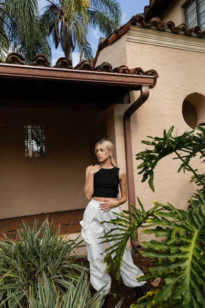 Junge Frau in trendigen Klamotten steht neben modernem Haus und grünen Pflanzen in Miami — Stockfoto