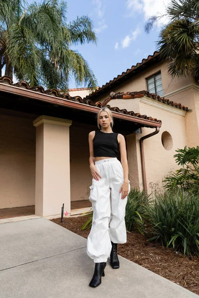 Full length of young woman in trendy clothes and boots standing near modern house and green plants in Miami — Stock Photo