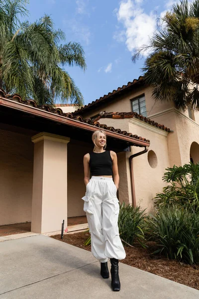 Piena lunghezza di donna felice in abiti alla moda in piedi con le mani in tasca vicino alla casa moderna a Miami — Stock Photo