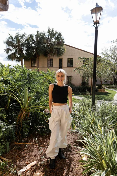 Повний зріст позитивної жінки в модному одязі стоїть з руками в кишенях біля сучасного будинку в Маямі. — Stock Photo