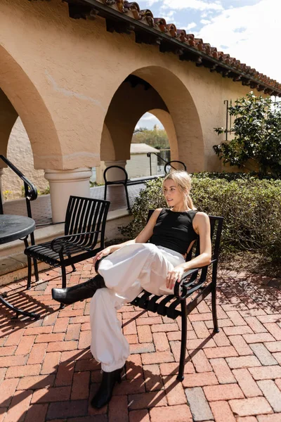Full length of blonde woman in trendy clothes sitting on patio chair near modern house in Miami — Stock Photo