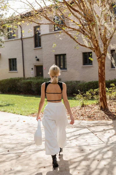 Vista trasera de mujer rubia con bolso caminando cerca de casa en Miami - foto de stock