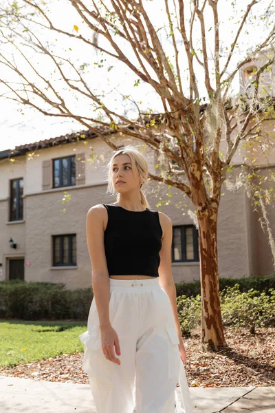 Blonde woman in black tank top looking away while standing near house in Miami — Stock Photo