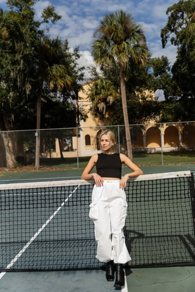 Full length of blonde woman in tank top and white cargo pants leaning on net in tennis court — Stock Photo